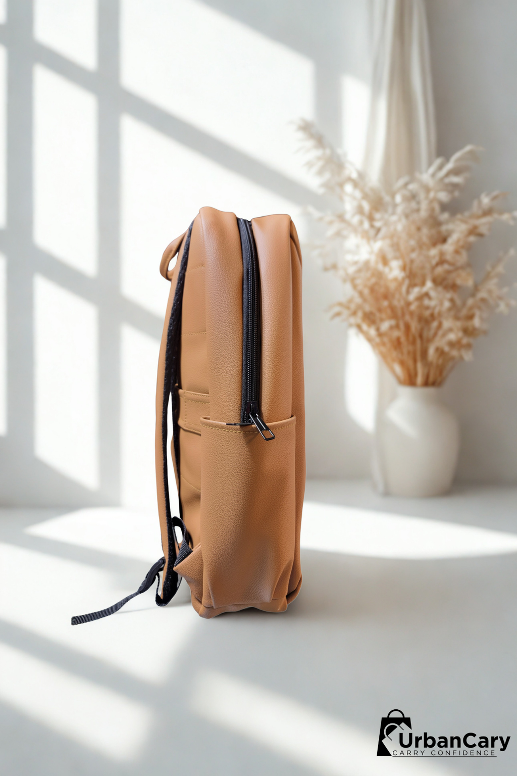 A tan leather backpack with black zippers and straps, standing upright on a white surface in a brightly lit room with window shadows. A white vase with dried pampas grass is in the background. Mens Leather Backpack