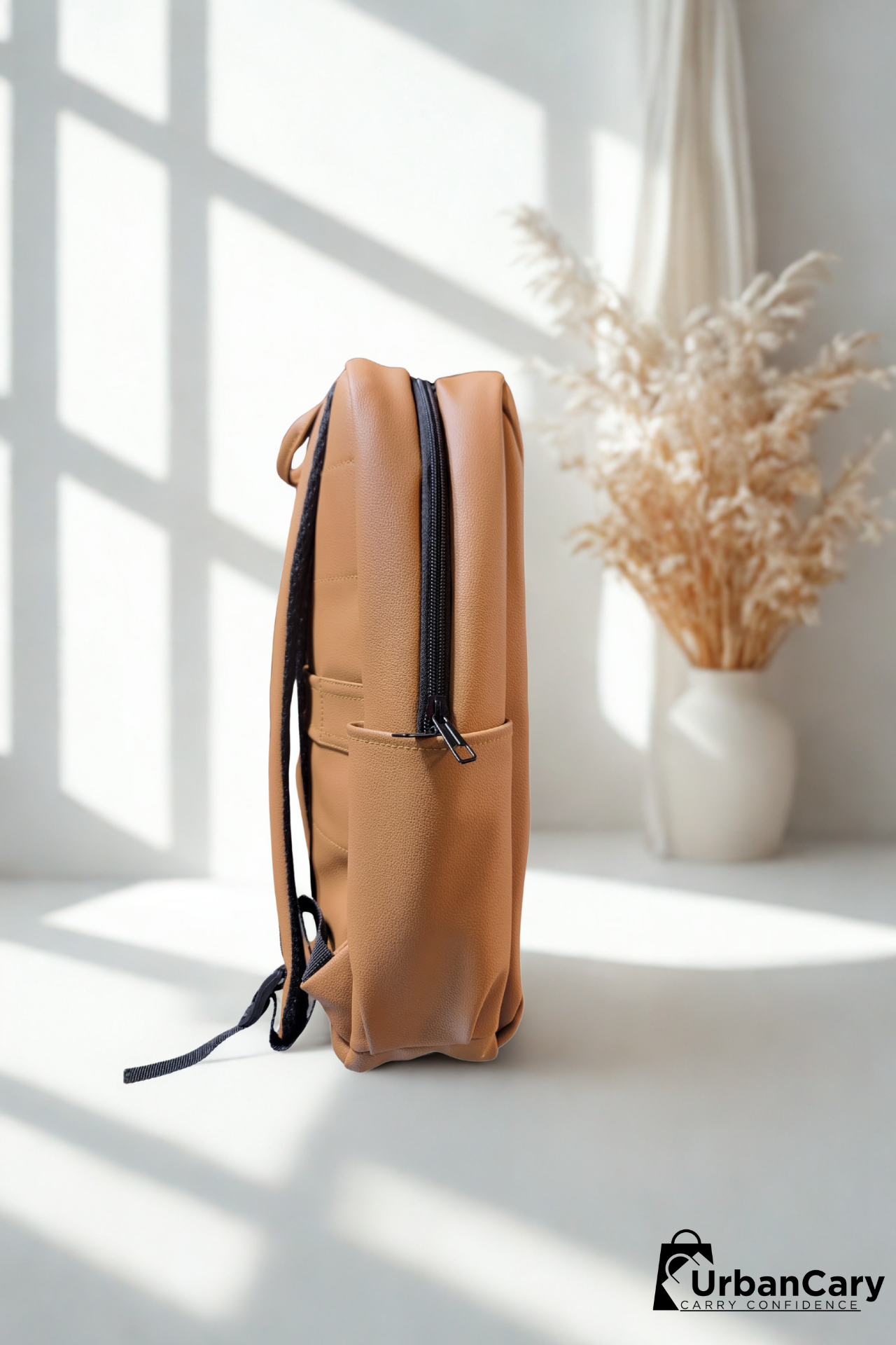 A tan leather backpack with black zippers and straps, standing upright on a white surface in a brightly lit room with window shadows. A white vase with dried pampas grass is in the background. Mens Leather Backpack