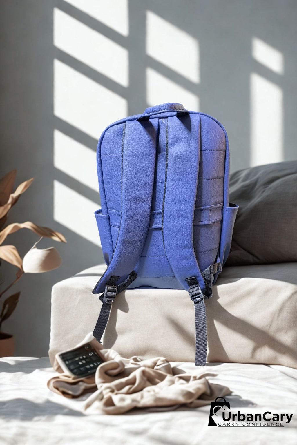 A blue backpack shown from the back, resting on a beige cushion in a sunlit room with window shadows. A beige towel and a black calculator are on the bed beside it, and the UrbanCary logo appears in the bottom right corner. Mens Leather Backpack