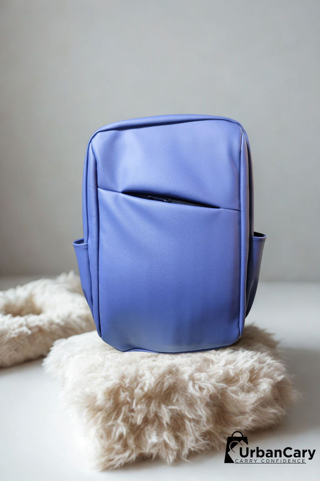 A blue backpack with a front slip pocket, displayed on a fluffy cream faux fur rug against a neutral gray background. The UrbanCary logo is visible in the bottom right corner. Mens Leather Backpack