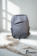 A gray minimalist backpack with a front zippered pocket, styled on a white surface with a cream-colored knit blanket under soft window shadows. The UrbanCary logo is visible in the bottom right corner. Mens Leather Backpack