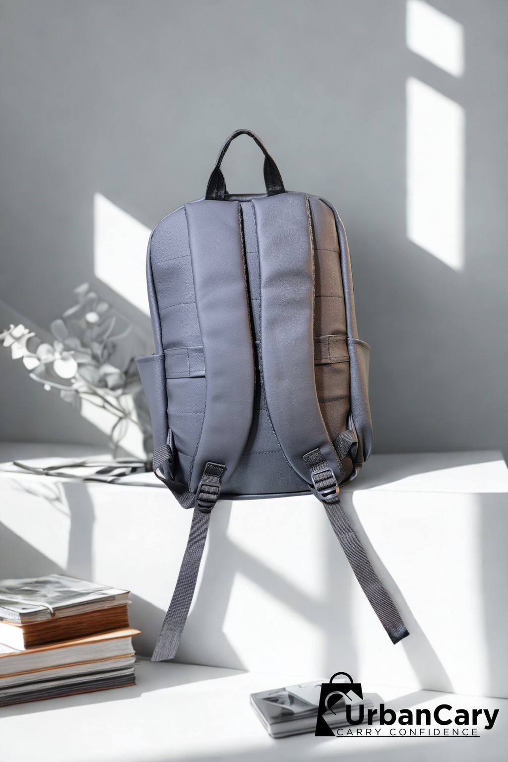A gray backpack with black straps and handles, shown from the back on a white platform in a minimalist room with window shadows. Books and a vase are on the left, and an UrbanCary logo is in the bottom right corner. Mens Leather Backpack