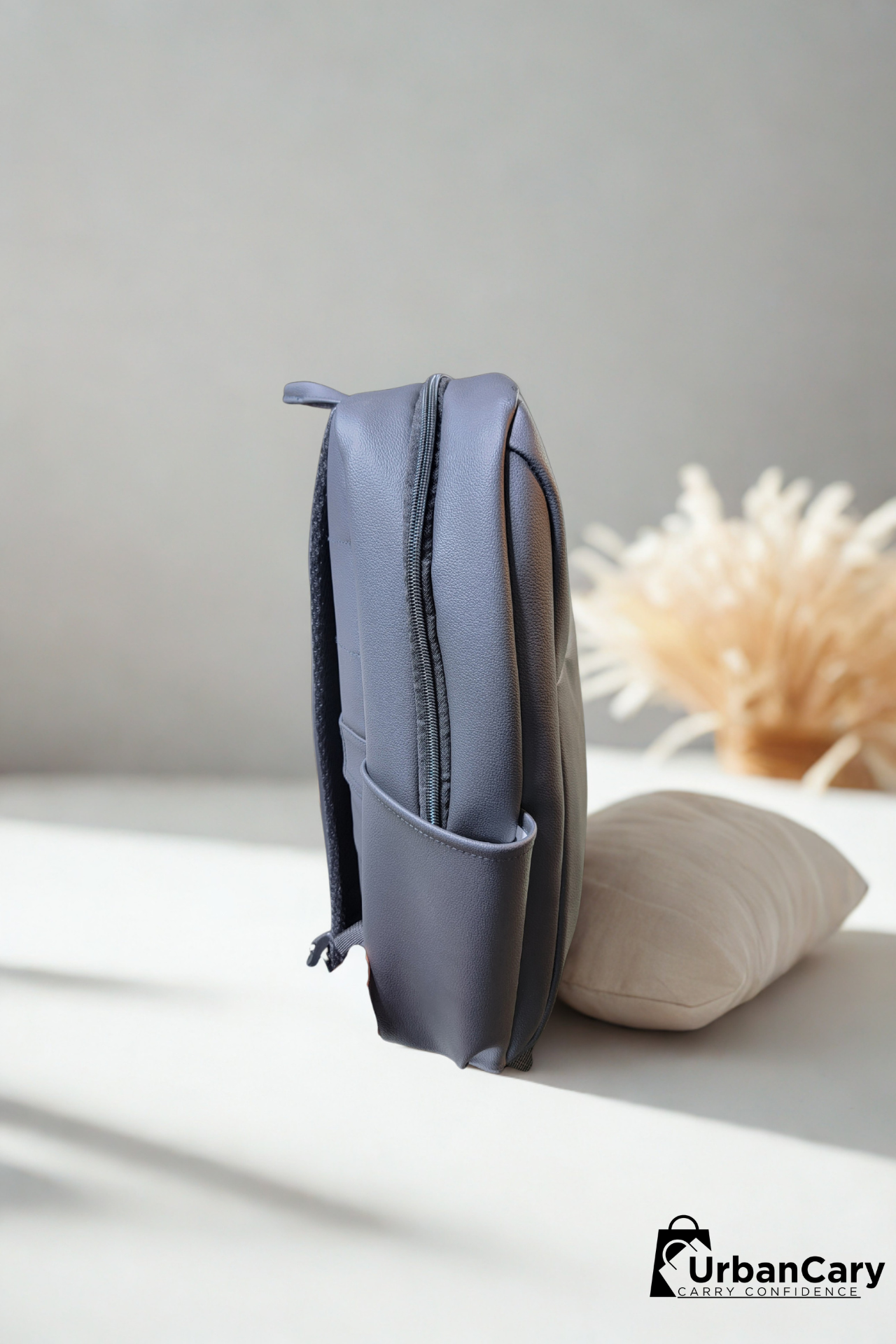 A slate blue-gray leather backpack standing upright on a white surface next to a beige pillow, with a soft-focus dried floral arrangement in the background. The UrbanCary logo is visible in the bottom right corner. Mens Leather Backpack